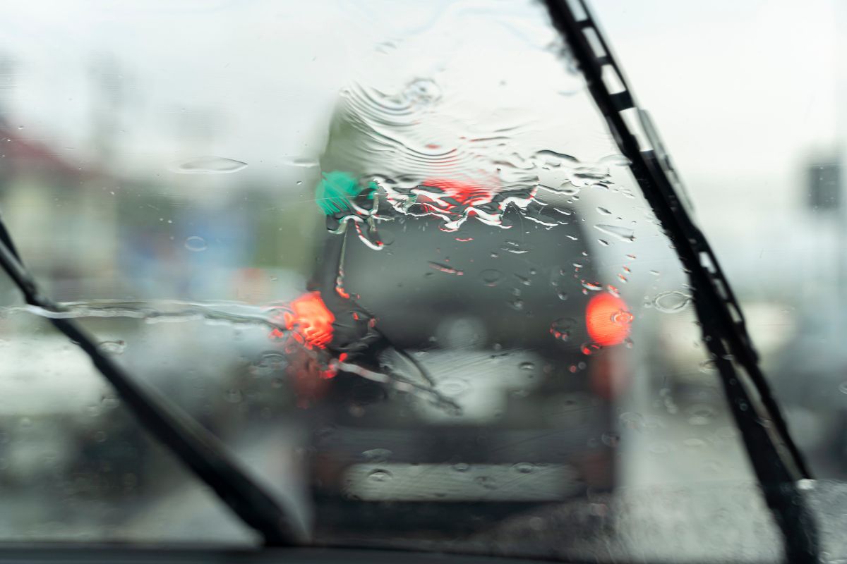 Rain-covered windshield from inside a vehicle, showing wipers in motion and traffic lights blurred by water—illustrating a potential leak or visibility issue.