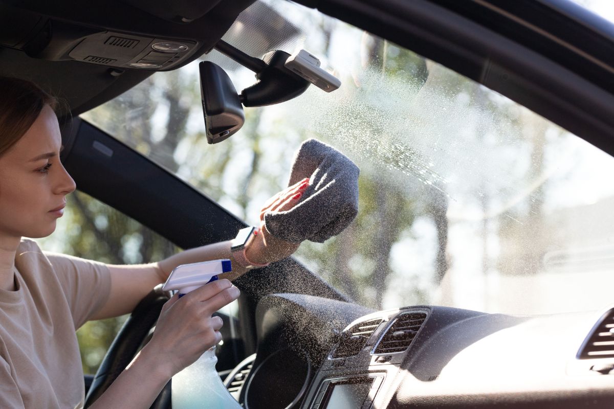 Driver cleaning a windshield with a microfiber towel and cleaner, showing the best way to clean windshield without streaks – clear glass with sun reflecting off it.