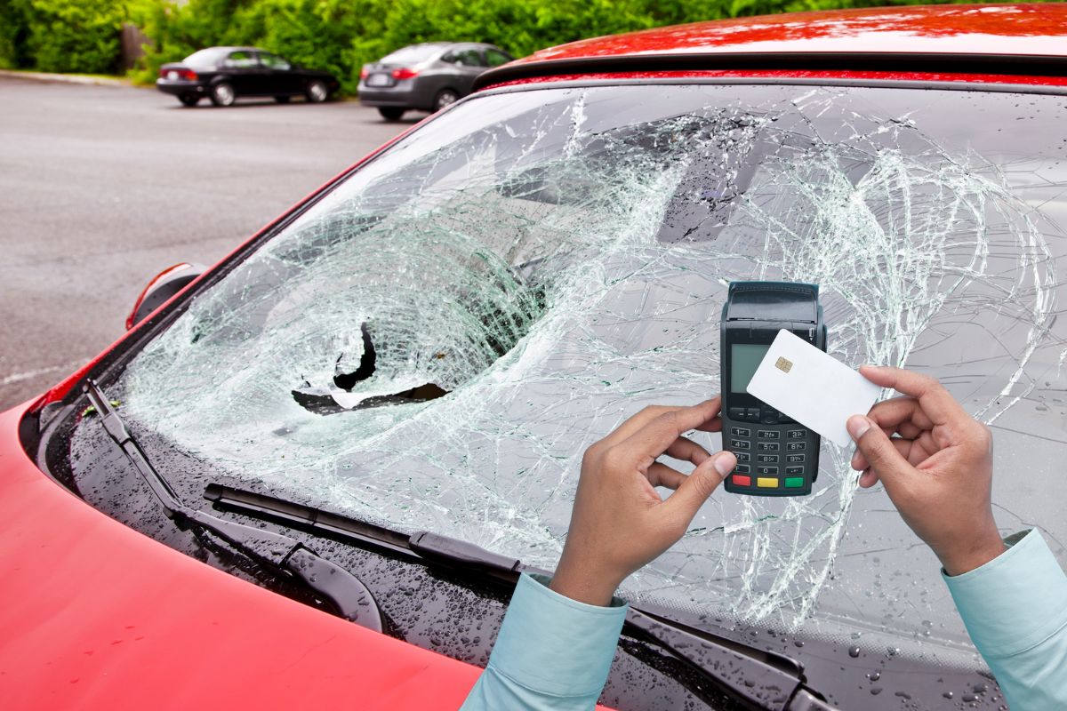 A person holding a payment card and machine in front of a severely cracked and shattered car windshield, symbolizing paying out-of-pocket for windshield repairs.