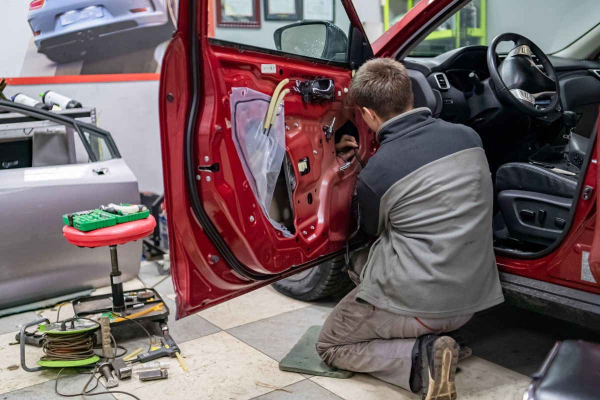 A technician kneeling beside a red vehicle with its interior door panel removed while repairing a window mechanism inside an auto repair shop.