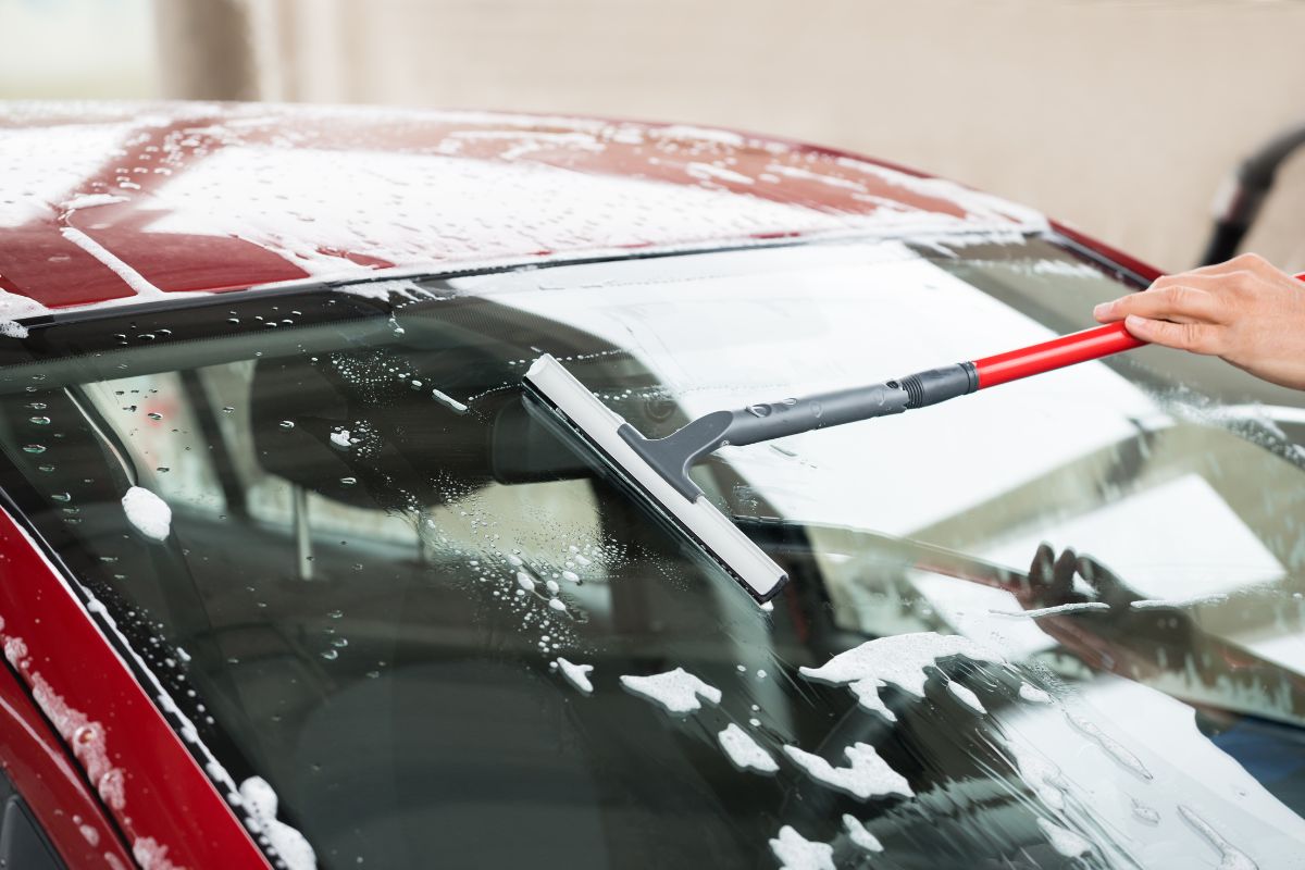 A person washing a car windshield with a long-handled squeegee, with soap and water covering the glass of a red vehicle.