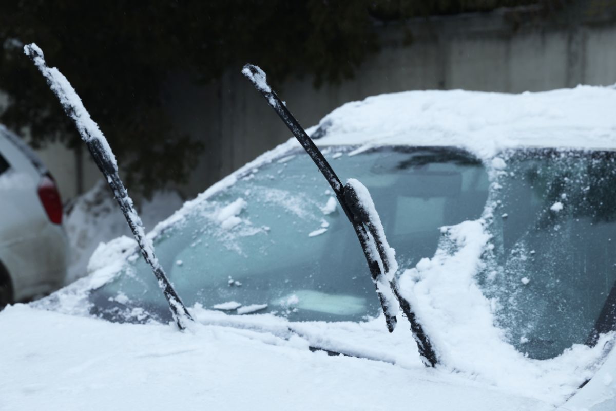 Car windshield covered in snow with raised wipers, showing how winter conditions in Wisconsin can cause windshield cracks to spread faster.